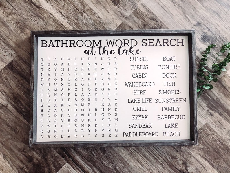 Puede incluir: Un letrero de madera blanco con marco negro y el texto "Bathroom Word Search at the Lake" impreso en negro. El letrero presenta un crucigrama de palabras con palabras relacionadas con el lago, como "barco", "caba&ntilde;a", "pescado", "parrilla", "kayak", "lago", "tabla de remo", "atardecer", "surf", "tubing" y "wakeboard".