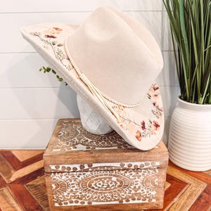May include: A cream-colored cowboy hat with floral embroidery and a cream-colored tassel. The hat rests on a white vase atop a wooden box with carved white designs. The box sits on a wooden table with a geometric pattern.