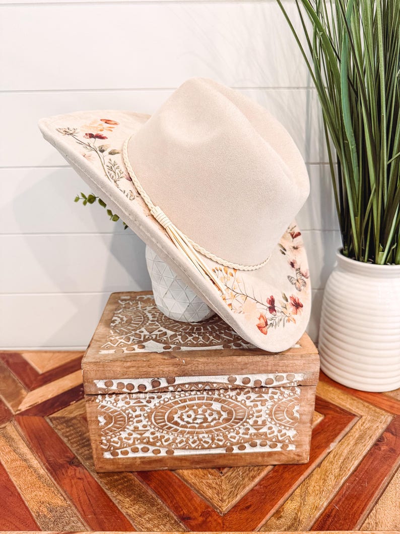 May include: A cream-colored cowboy hat with floral embroidery and a cream-colored tassel. The hat rests on a white vase atop a wooden box with carved white designs. The box sits on a wooden table with a geometric pattern.