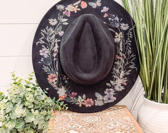May include: A black wide-brimmed hat with a floral print design. The hat has a black band and a black crown. The floral print features butterflies, leaves, and flowers in shades of pink, purple, and white.