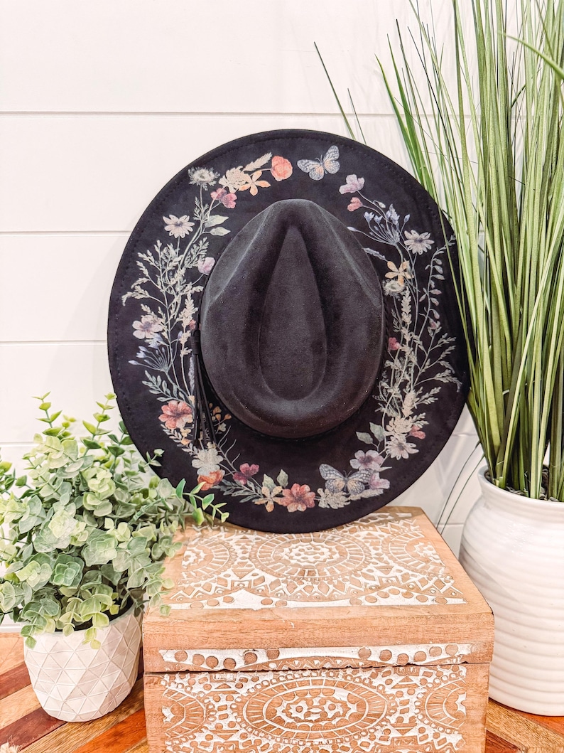 Op de afbeelding: Een zwarte fedora-hoed met een brede rand, versierd met een bloemen- en vlinderpatroon in roze, perzik en lavendel. De hoed heeft een donkergrijze kroon en een kwast. De hoed ligt op een houten kist.