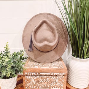 May include: A taupe-colored fedora hat with a heart-shaped crown and a decorative tassel. The hat sits atop a wooden box with white carved designs. The box is on a table with a geometric pattern.