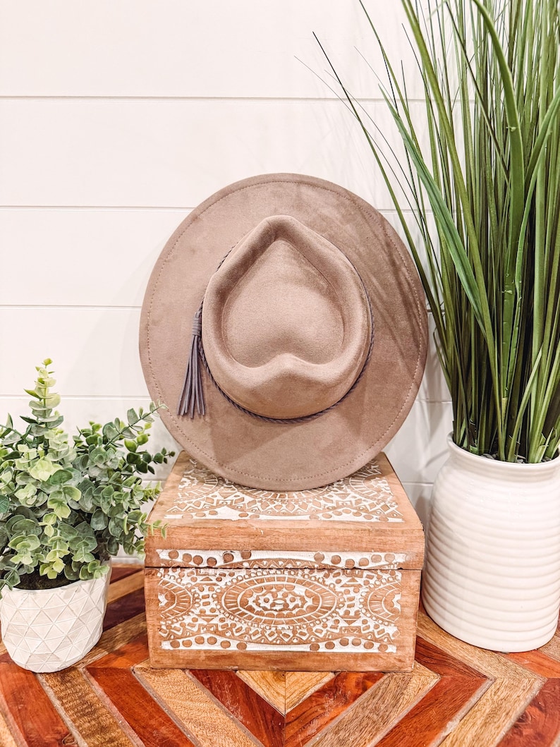 May include: A taupe-colored fedora hat with a heart-shaped crown and a decorative tassel. The hat sits atop a wooden box with white carved designs. The box is on a table with a geometric pattern.