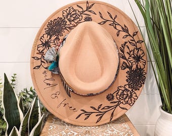 Puede incluir: Un sombrero fedora de fieltro marrón con un diseño floral negro y el texto "wild & free" en el borde. El sombrero está decorado con una pluma turquesa y negra.