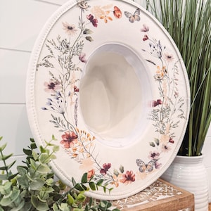 Op de afbeelding: Een cr&egrave;mekleurige fedora hoed met een bloemen- en vlinderontwerp. De hoed heeft een brede rand en een cr&egrave;mekleurig interieur. Het bloemmotief bevat bloemen in tinten rood, roze en oranje. Een stijlvol accessoire.