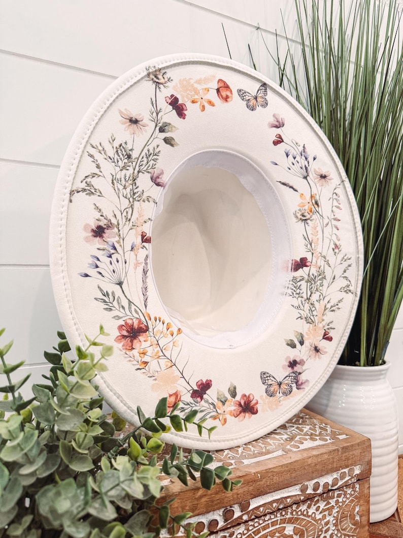 Op de afbeelding: Een cr&egrave;mekleurige fedora hoed met een bloemen- en vlinderontwerp. De hoed heeft een brede rand en een cr&egrave;mekleurig interieur. Het bloemmotief bevat bloemen in tinten rood, roze en oranje. Een stijlvol accessoire.