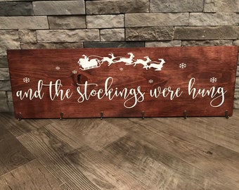 May include: A wooden sign with the text "and the stockings were hung" in white paint. The sign features a silhouette of Santa Claus in his sleigh with reindeer flying above. The sign has hooks for hanging stockings.