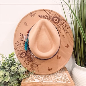 May include: A tan fedora-style hat with a wide brim, decorated with etched sunflowers and butterflies. The hat features a decorative band and a small tassel. The hat is resting on a wooden box with carved designs.