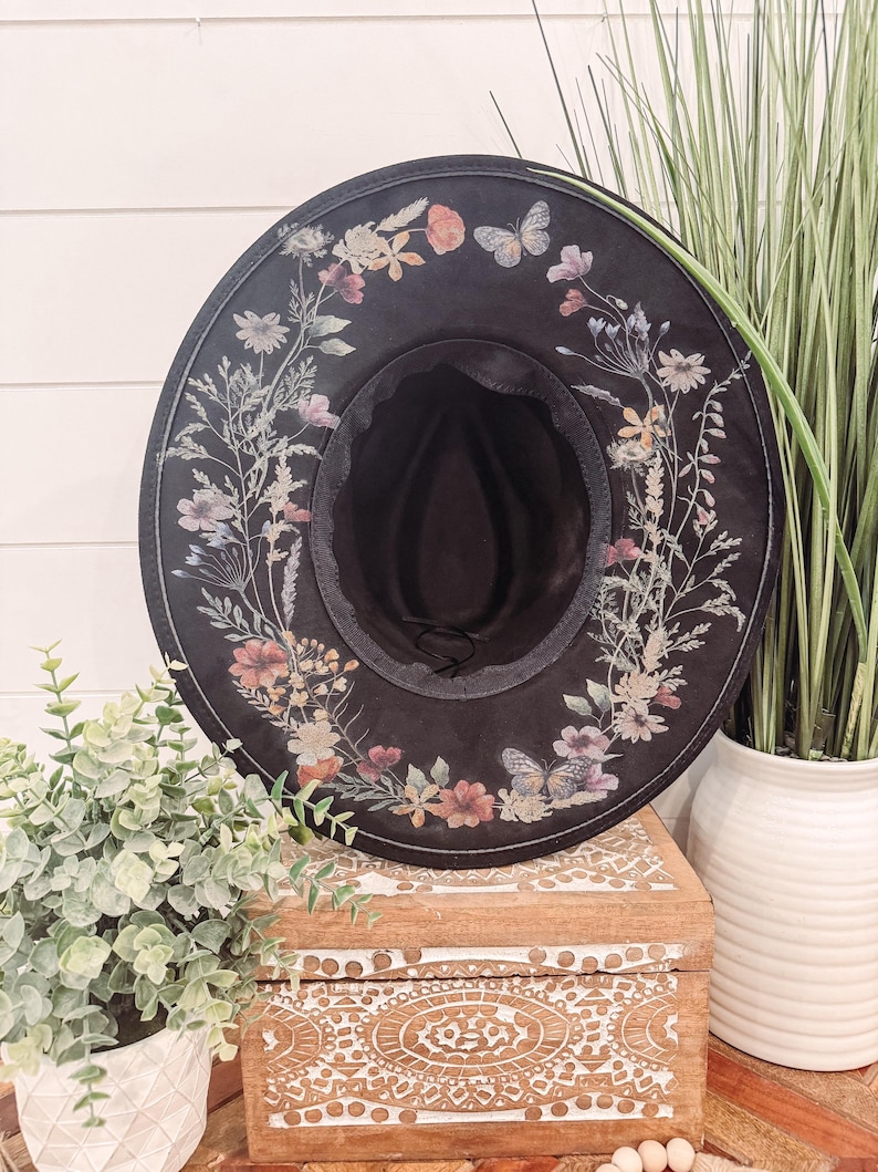 May include: A wide-brimmed black hat with a floral print design. The hat has a black band around the crown and a floral print around the brim. The hat is sitting on a wooden box with a white and brown design.