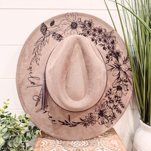 May include: A brown felt fedora hat with a black floral design and the text "wild one" on the brim.