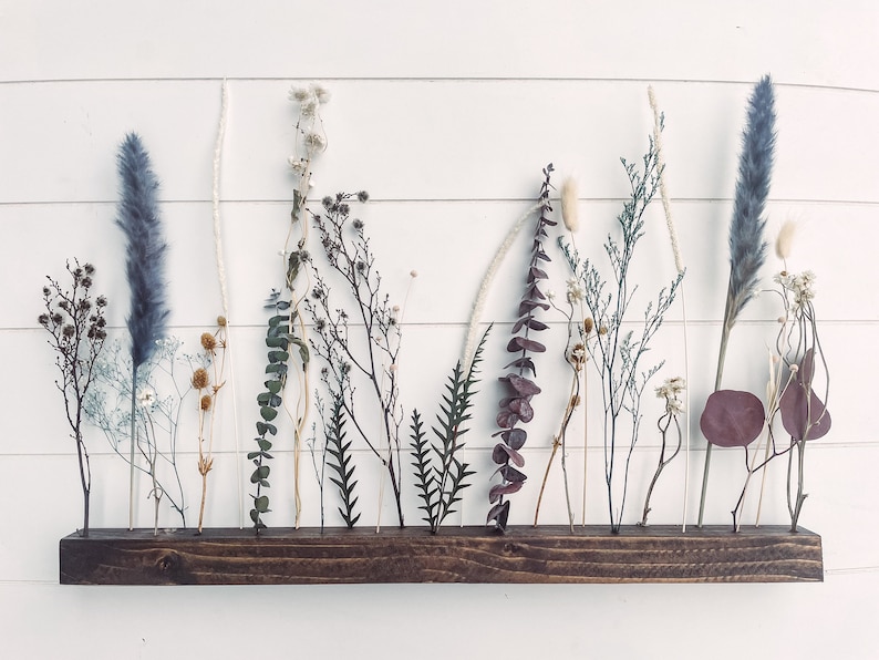 K&ouml;nnte beinhalten: Eine Wandhalterung aus Holz mit getrockneten Blumen und Gr&auml;sern in Glasr&ouml;hrchen. Die Anzeige zeigt eine Vielzahl von Farben und Texturen, darunter Blau, Lila, Wei&szlig; und Braun.