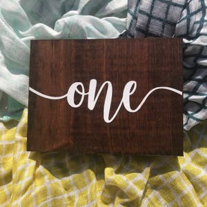May include: A wooden sign with the word "one" written in white cursive script on a dark brown background.