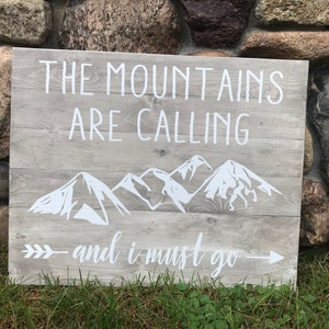 Puede incluir: Un letrero de madera con el texto "The Mountains Are Calling - and I Must Go" pintado en blanco. El letrero tambi&eacute;n presenta una silueta blanca de monta&ntilde;as y una flecha.