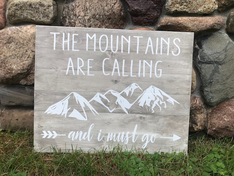 Puede incluir: Un letrero de madera con el texto "The Mountains Are Calling - and I Must Go" pintado en blanco. El letrero tambi&eacute;n presenta una silueta blanca de monta&ntilde;as y una flecha.