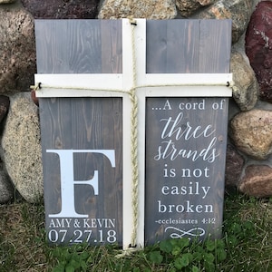 May include: A gray wooden sign with a white cross and the text "...A cord of three strands is not easily broken -ecclesiastes 4:12". The sign also has a white letter "F" and the text "AMY & KEVIN 07.27.18".