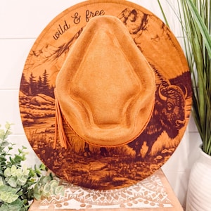 May include: A tan fedora-style hat with a wide brim featuring a landscape design and a bison illustration. The hat has the words "wild & free" printed on the brim. A tan tassel hangs from the side of the hat.