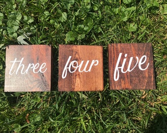 May include: Three wooden signs with the words "three", "four", and "five" painted in white on a dark brown background.