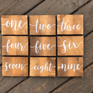 May include: Nine wooden signs with the numbers one through nine written in white cursive font on a brown wooden background.