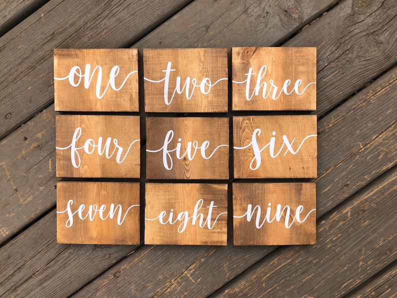 May include: Nine wooden signs with the numbers one through nine written in white cursive font on a brown wooden background.