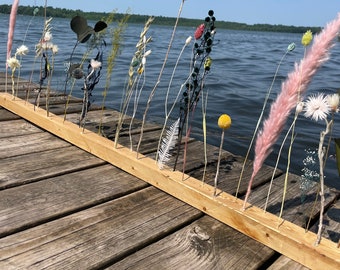 Könnte beinhalten: Ein Holzbrett mit getrockneten Blumen und Gräsern, die in einer Reihe angeordnet sind. Das Brett liegt auf einem Holzsteg mit einem See im Hintergrund.
