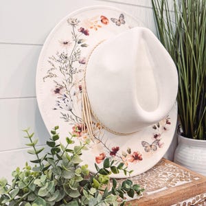 May include: A white wide-brimmed hat with a floral print and a butterfly design. The hat is sitting on a wooden surface with green plants.