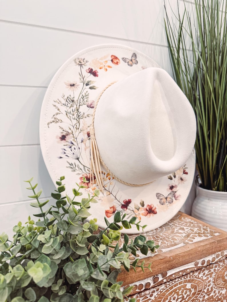 May include: A white wide-brimmed hat with a floral print and a butterfly design. The hat is sitting on a wooden surface with green plants.