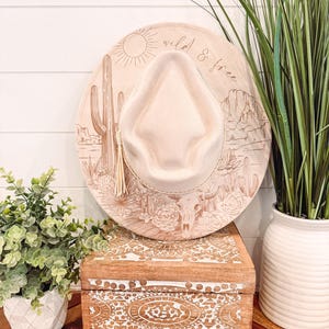 Op de afbeelding: Een beige fedora-hoed met een woestijnlandschapontwerp en de woorden "wild & free" gegraveerd op de rand. De hoed heeft een decoratieve kwast en wordt getoond op een houten kist met een gesneden ontwerp.
