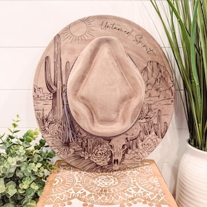 Op de afbeelding: Een beige fedora-hoed met een woestijnlandschapontwerp op de rand en de woorden "Untamed Spirit". De hoed wordt getoond op een houten kist met een wit gesneden ontwerp. De hoed heeft een kwast.