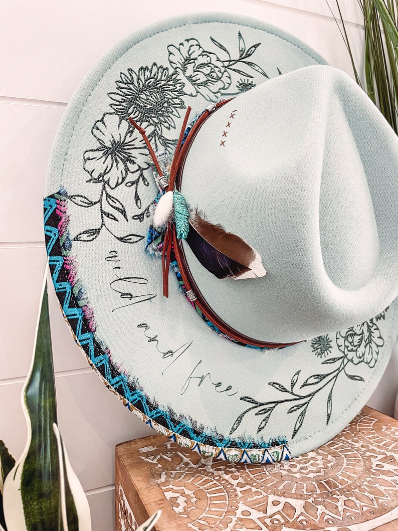 May include: A light blue felt hat with a black floral design and the words "wild and free" written on the brim. The hat has a brown leather band and a blue and black patterned trim.