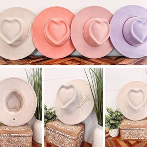 May include: A selection of wide-brimmed hats in pastel shades: beige, coral, pink, and lavender. Each hat has a heart-shaped crown and a decorative band. The hats are displayed on a wooden surface, highlighting their design and style. The hats are suitable for various occasions.