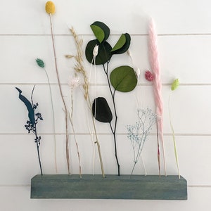 Pu&ograve; includere: Un ripiano in legno verde-azzurro con fiori secchi e verde. Il ripiano ha pi&ugrave; fori per tenere i gambi. I fiori includono un'erba della pampa rosa, una palla di billy gialla e una variet&agrave; di erbe e foglie secche.