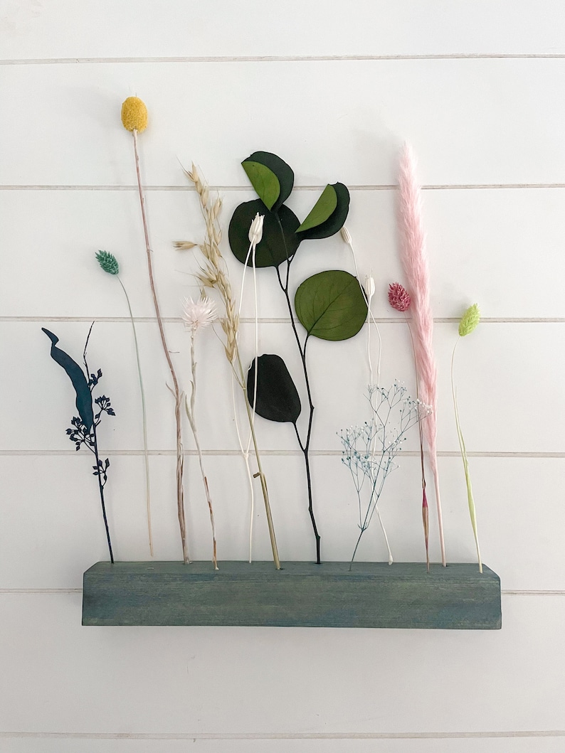 Pu&ograve; includere: Un ripiano in legno verde-azzurro con fiori secchi e verde. Il ripiano ha pi&ugrave; fori per tenere i gambi. I fiori includono un'erba della pampa rosa, una palla di billy gialla e una variet&agrave; di erbe e foglie secche.