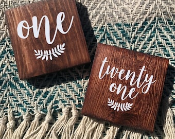 May include: Two wooden signs with white lettering. The first sign says "one" with a small leafy branch design. The second sign says "twenty one" with a small leafy branch design.