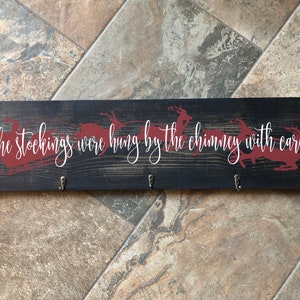 May include: A black wooden stocking holder with the text "The stockings were hung by the chimney with care..." and red reindeer silhouettes. The holder has three hooks for hanging stockings.