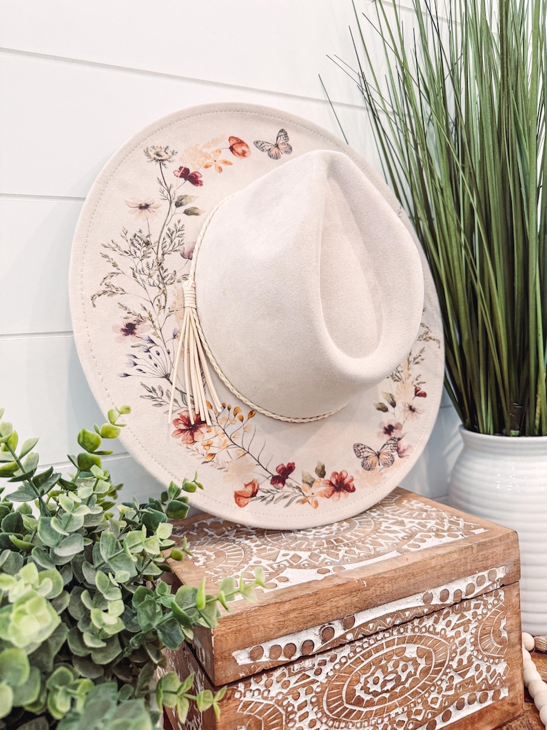 May include: A beige fedora hat with a floral and butterfly print. The hat is sitting on a wooden box with a carved design.