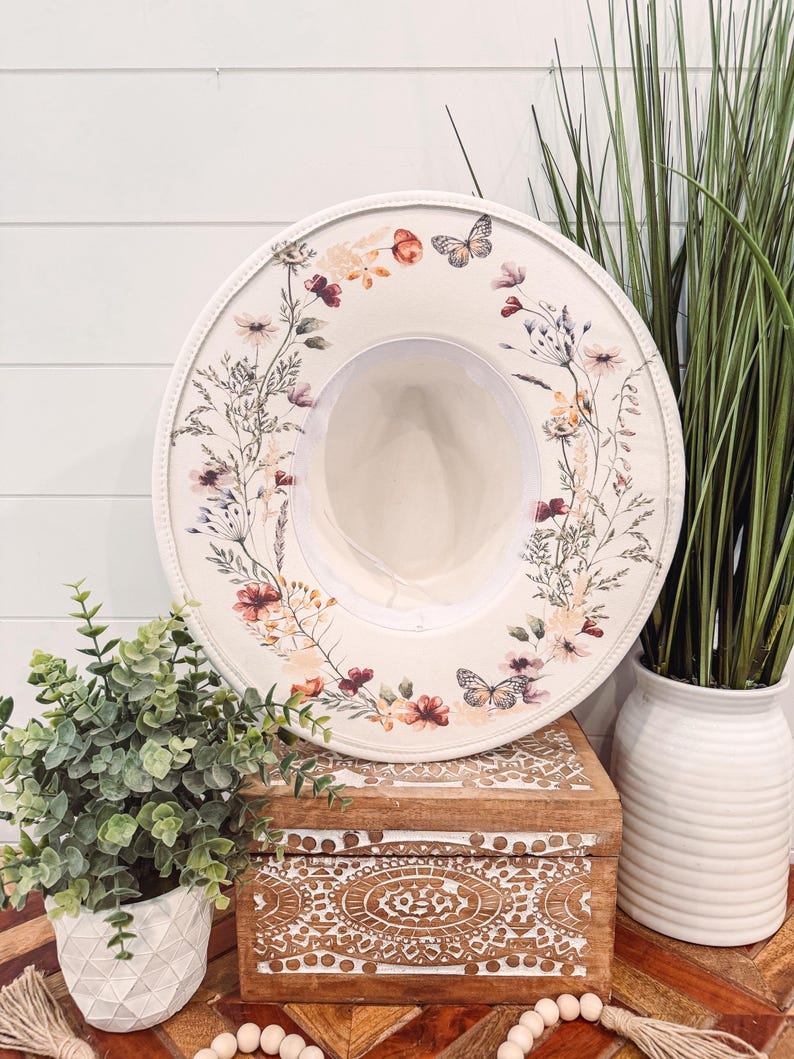May include: A white sun hat with a floral and butterfly design. The hat is sitting on a wooden box with a carved design.