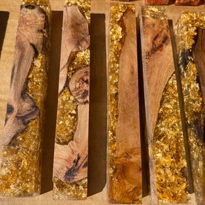 May include: Close-up of several rectangular wooden blocks encased in clear resin with gold leaf accents. The wood grain varies, showcasing natural patterns. The gold leaf adds a luxurious touch to the handcrafted pieces.