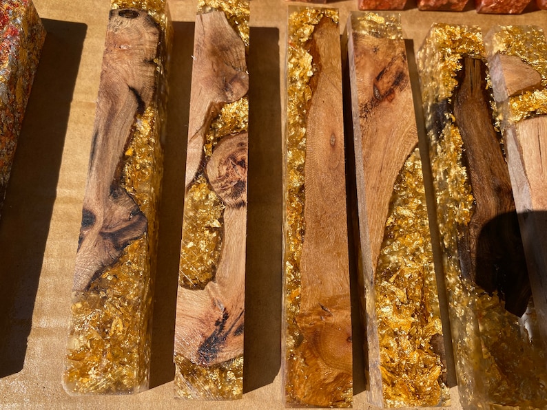 May include: Close-up of several rectangular wooden blocks encased in clear resin with gold leaf accents. The wood grain varies, showcasing natural patterns. The gold leaf adds a luxurious touch to the handcrafted pieces.