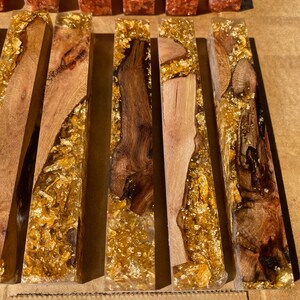 May include: Close-up of several rectangular wooden blocks encased in clear resin. The wood varies in color from light to dark brown, with gold leaf accents throughout. The blocks are arranged side by side.