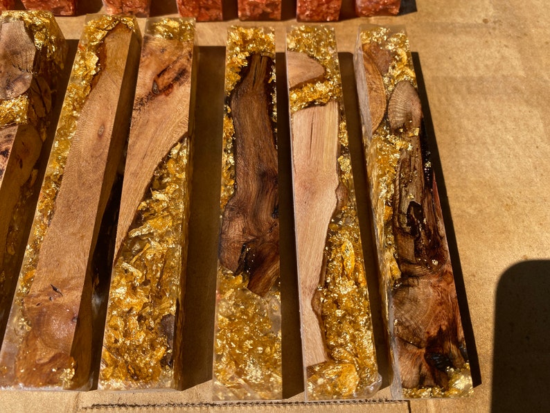 May include: Close-up of several rectangular wooden blocks encased in clear resin. The wood varies in color from light to dark brown, with gold leaf accents throughout. The blocks are arranged side by side.