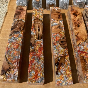 May include: Close-up of several rectangular wooden blocks with a clear resin finish. The blocks feature a mix of wood grain, metallic flakes in silver, gold, and copper, creating a textured, decorative appearance. These blocks could be used for crafting.