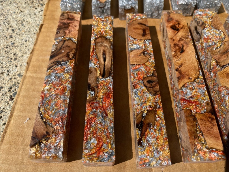May include: Close-up of several rectangular wooden blocks with a clear resin finish. The blocks feature a mix of wood grain, metallic flakes in silver, gold, and copper, creating a textured, decorative appearance. These blocks could be used for crafting.