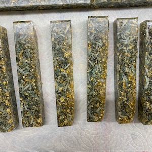 Può includere: Sette blocchi rettangolari in resina con banconote di carta verde e oro incorporate. I blocchi sono disposti in fila su una superficie bianca.