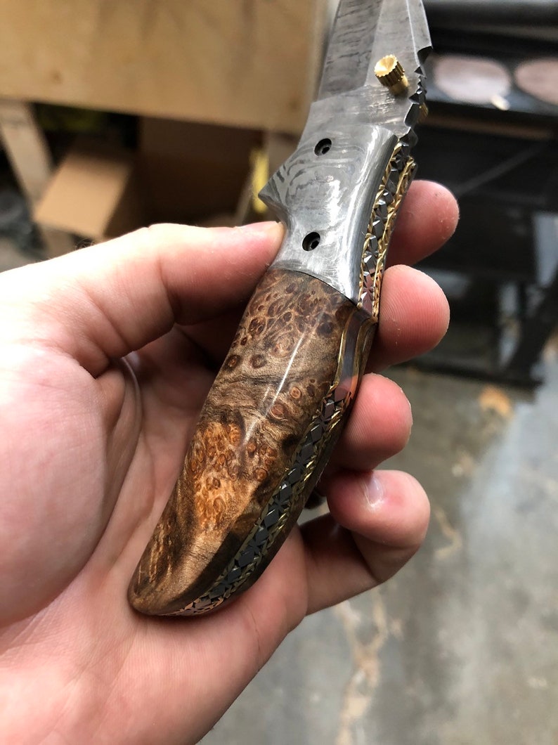 Pu&ograve; includere: Un coltello pieghevole su misura con manico in legno e un'intarsio in metallo decorativo. La lama &egrave; in acciaio a motivi e ha un perno in ottone.