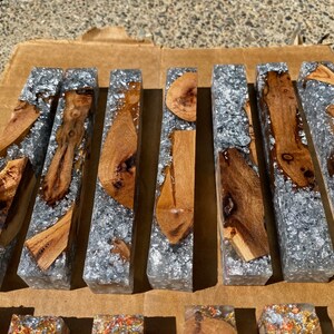 May include: A collection of rectangular resin blocks with embedded wood pieces and silver flakes. The wood varies in shape and grain, with light brown and dark brown tones. The resin is clear, and the silver flakes add a shimmering effect.
