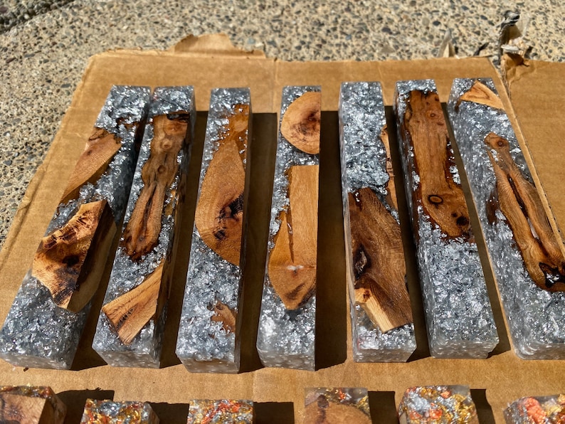 May include: A collection of rectangular resin blocks with embedded wood pieces and silver flakes. The wood varies in shape and grain, with light brown and dark brown tones. The resin is clear, and the silver flakes add a shimmering effect.