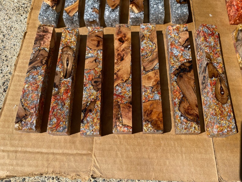 May include: A collection of rectangular resin blocks with wood and metallic inclusions. The wood pieces are brown and tan, while the metallic elements are silver, copper, and gold. The blocks are arranged on a brown surface.