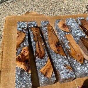 May include: Close-up of several rectangular resin blocks with embedded wood pieces and silver flakes. The blocks are arranged on a brown cardboard surface, showcasing the contrast between the natural wood and the shimmering resin.