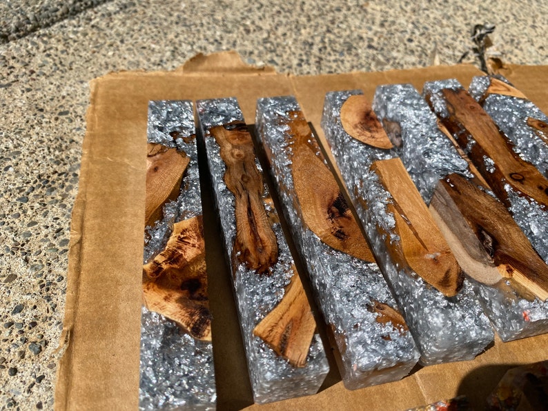 May include: Close-up of several rectangular resin blocks with embedded wood pieces and silver flakes. The blocks are arranged on a brown cardboard surface, showcasing the contrast between the natural wood and the shimmering resin.