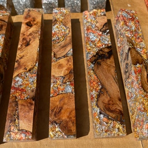 May include: Close-up of several rectangular wooden blocks with a clear resin finish. The wood has a natural grain pattern and is interspersed with silver, copper, and gold metallic flakes. The blocks are arranged side by side.